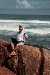 Cyfrowy nomada, człowiek pracujący z laptopem i smartfonem zdalnie na tle oceanu i skał siedzący na wybrzeżu.