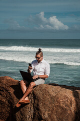 Cyfrowy nomada, człowiek pracujący z laptopem i smartfonem zdalnie na tle oceanu i skał siedzący na wybrzeżu. - obrazy, fototapety, plakaty