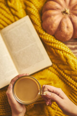 Fragrant coffee in women's hands, a book of stories on knitted sweater. Top view of a romantic background. Ripe round pumpkin. Autumn composition for greeting cards and invitations. Fall still life.