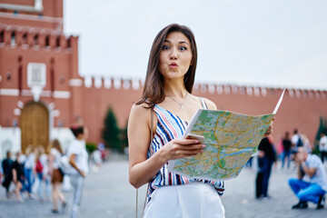 Half length portrait of attractive hipster girl with duck face and location paper in hands posing in historic center, Asian woman with travel map showing kiss while looking at camera during vacations