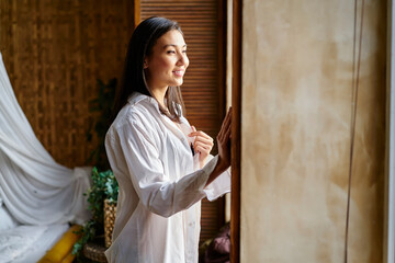 Happy Asian woman dressed in white shirt looking in hotel window and smiling during Sunday morning,...