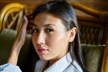 Portrait of attractive female model with natural makeup looking at camera during photo session indoors, close up of young Asian woman with brunette hair posing during leisure pastime inside