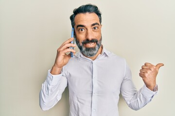 Middle age man with beard and grey hair having conversation talking on the smartphone pointing thumb up to the side smiling happy with open mouth