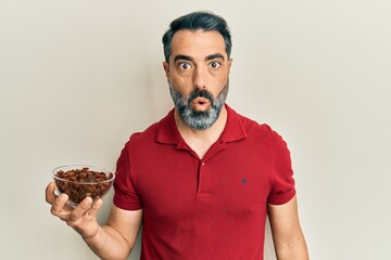 Middle age man with beard and grey hair holding raisins bowl scared and amazed with open mouth for surprise, disbelief face