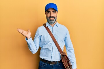 Middle age man with beard and grey hair wearing delivery courier cap smiling cheerful presenting and pointing with palm of hand looking at the camera.