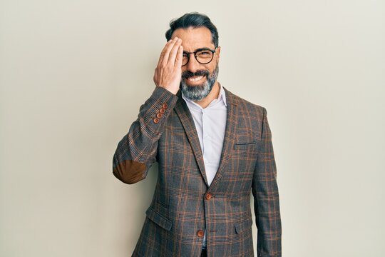 Middle age man with beard and grey hair wearing business jacket and glasses covering one eye with hand, confident smile on face and surprise emotion.