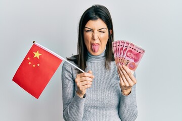 Young hispanic woman holding china flag and yuan banknotes sticking tongue out happy with funny...