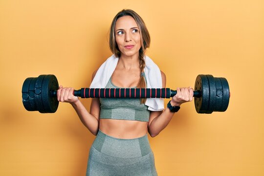 Beautiful hispanic woman wearing sportswear using dumbbells smiling looking to the side and staring away thinking.