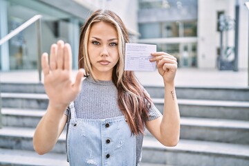 Young caucasian woman holding covid record card with open hand doing stop sign with serious and...