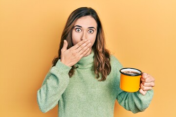 Young hispanic girl drinking a cup of coffee covering mouth with hand, shocked and afraid for mistake. surprised expression