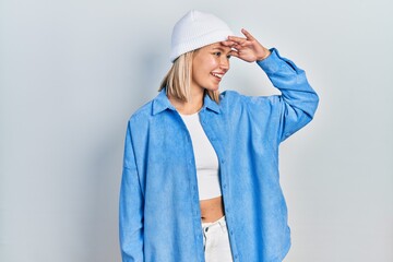 Beautiful blonde woman wearing wool hat very happy and smiling looking far away with hand over head. searching concept.