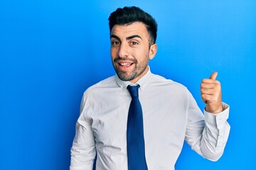 Young hispanic man wearing business clothes smiling with happy face looking and pointing to the side with thumb up.