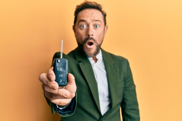 Middle age businessman holding key of new car scared and amazed with open mouth for surprise,...