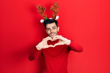 Young hispanic man wearing cute christmas reindeer horns smiling in love doing heart symbol shape with hands. romantic concept.