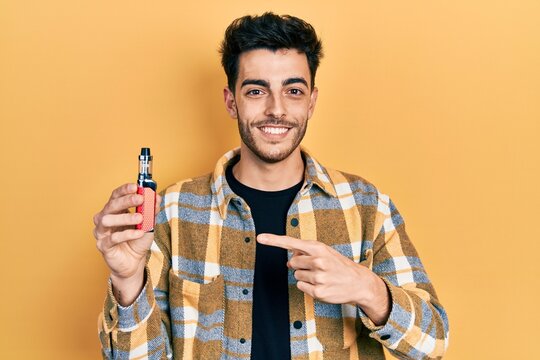 Young hispanic man holding electronic cigarette smiling happy pointing with hand and finger