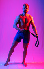 muscular man in blue shorts shakes muscles or poses with an elastic band for fitness. multi-colored light. purple and crimson highlights on face and background