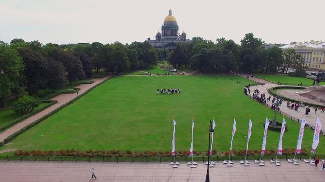 People walk along the green lawn near St. Isaac's Cathedral in the center of St. Petersburg, Russia. Quadcopter flies back and tourists, monuments, cars, the Neva River and boats appear. 4K Aerial