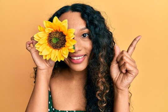 Young latin woman holding sunflower over eye smiling happy pointing with hand and finger to the side