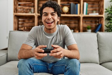 Young hispanic man playing video game sitting on the sofa at home.
