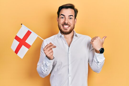 Handsome caucasian man with beard holding england flag pointing thumb up to the side smiling happy with open mouth