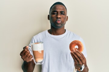 Young african american man eating doughnut and drinking coffee puffing cheeks with funny face. mouth inflated with air, catching air.