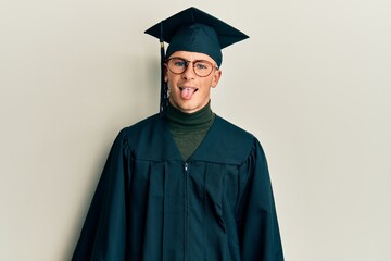 Young caucasian man wearing graduation cap and ceremony robe sticking tongue out happy with funny...