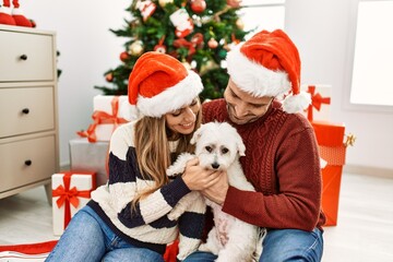 Young hispanic couple wearing christmas hat sitting on the floor with dog at home.