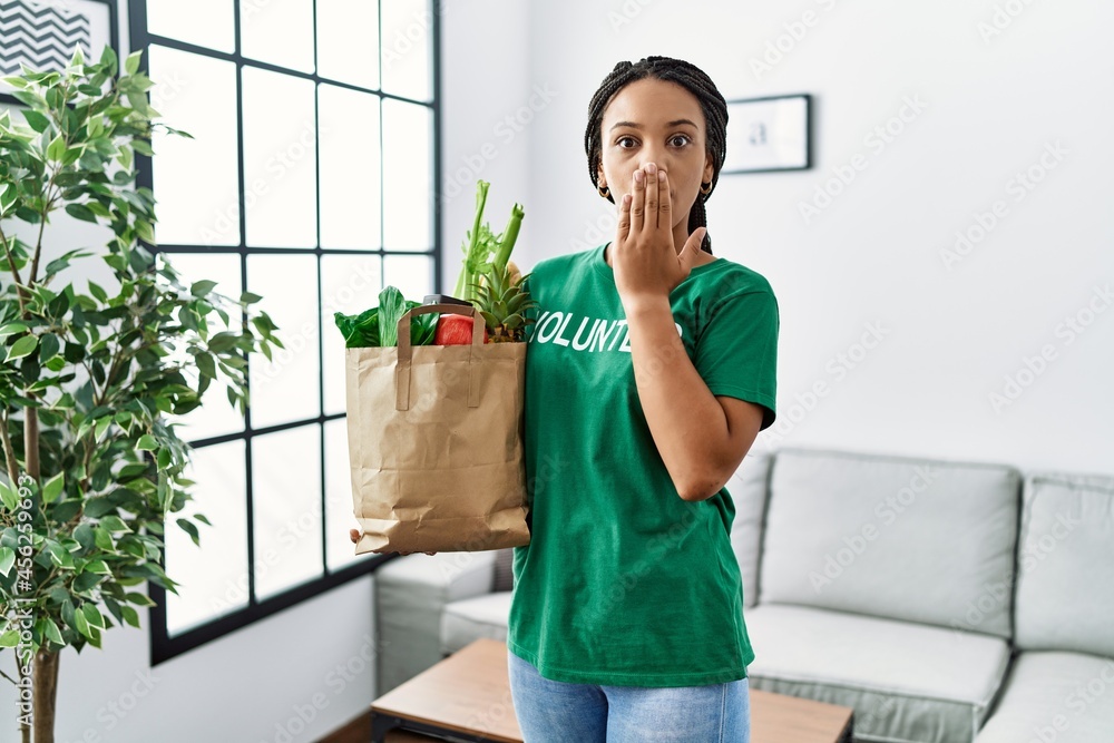 Wall mural Young african american woman wearing volunteer t shirt holding bag of groceries covering mouth with hand, shocked and afraid for mistake. surprised expression - Wall murals