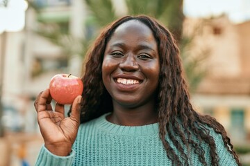 Young african woman smiling happy holding fresh red apple at the city