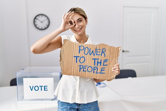 Young blonde woman at political election holding power to the people banner smiling happy doing ok sign with hand on eye looking through fingers