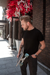 Handsome blode tattoed man skateboarder with longboard standing near city street brick wall on background of red floral bush looking thoughtful wears casual black denim outfit smart watch.