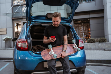 Handsome blonde male skater holding longboard sitting on car trunk parking using smartphone. Urban habits. Extreme sport concept. Communication concept. City life concept.  Outdoors leisure concept.