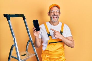 Handsome middle age man with grey hair standing by ladder showing smartphone smiling and laughing hard out loud because funny crazy joke.