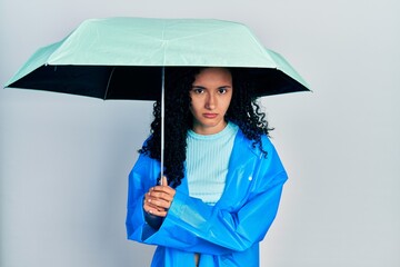 Young hispanic woman with curly hair wearing a raincoat and umbrella skeptic and nervous, frowning upset because of problem. negative person.