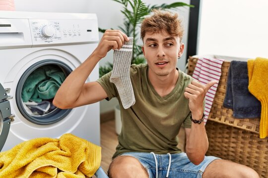 Young caucasian man doing laundry holding sock pointing thumb up to the side smiling happy with open mouth