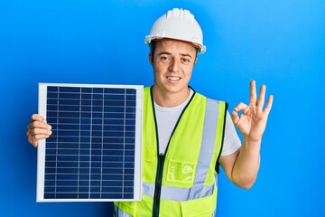 Handsome young man holding photovoltaic solar panel doing ok sign with fingers, smiling friendly gesturing excellent symbol