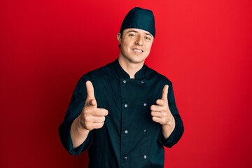 Handsome young man wearing professional cook uniform and hat pointing fingers to camera with happy...
