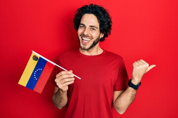 Handsome hispanic man holding venezuelan flag pointing thumb up to the side smiling happy with open mouth