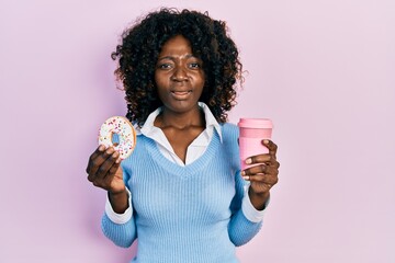 Young african american woman eating doughnut and drinking coffee clueless and confused expression....