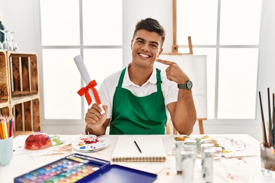 Young hispanic man at art studio holding degree smiling cheerful showing and pointing with fingers teeth and mouth. dental health concept.