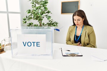 Young brunette woman sitting at election table with voting ballot with hand on stomach because...