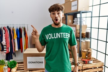 Young arab man wearing volunteer t shirt at donations stand surprised pointing with finger to the side, open mouth amazed expression.