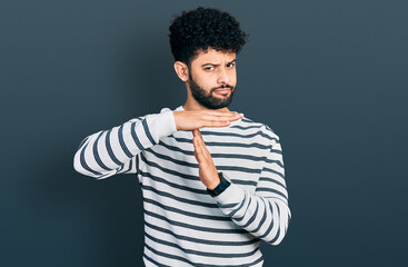Young arab man with beard wearing casual striped sweater doing time out gesture with hands,...