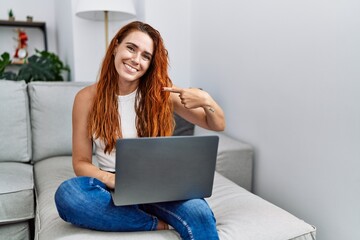 Young redhead woman using laptop at home pointing finger to one self smiling happy and proud