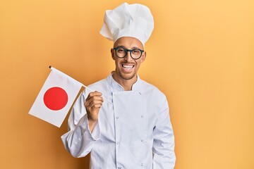 Bald man with beard wearing professional cook uniform holding japan flag looking positive and happy standing and smiling with a confident smile showing teeth