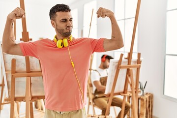 Young hispanic man at art studio showing arms muscles smiling proud. fitness concept.