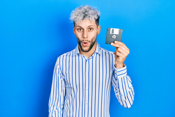 Young hispanic man with modern dyed hair holding floppy disk scared and amazed with open mouth for...