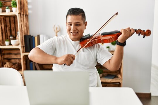 Young hispanic man having online violin lesson using laptop sitting on the table at home.