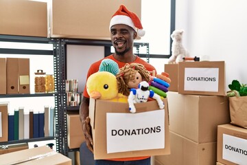 Young african man wearing volunteer t shirt and christmas hat holding donations winking looking at...