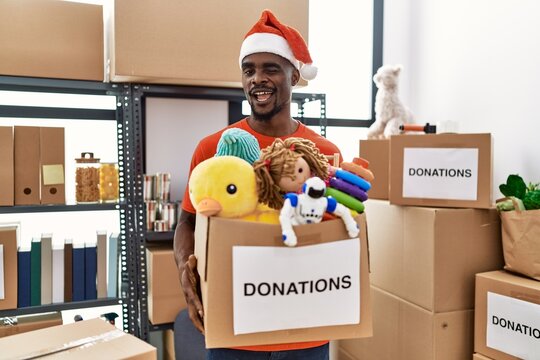 Young african man wearing volunteer t shirt and christmas hat holding donations winking looking at the camera with sexy expression, cheerful and happy face.